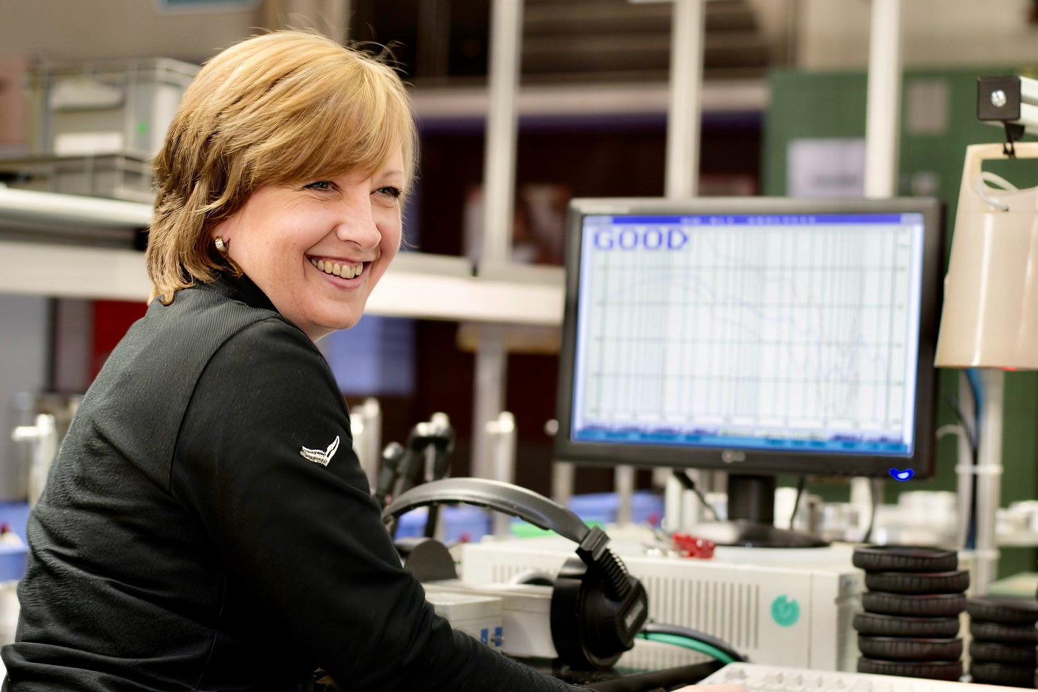 A friendly German Maestro technician smiles while performing a quality control test on a pair of headphones at her workstation. The computer monitor in front of her displays a graph and the word "GOOD," indicating a successful audio test.
