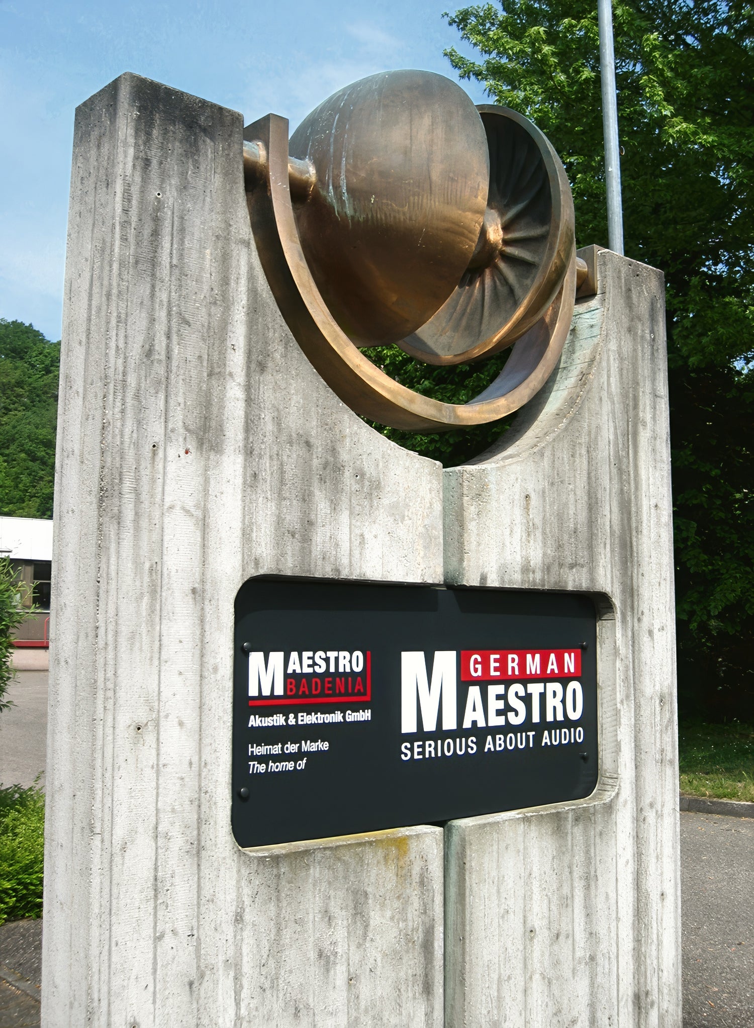 A modern concrete monument at the entrance of the German Maestro headquarters, topped with a large, stylized sculpture of brass headphones. A black plaque below the sculpture features the "Maestro Badenia" and "German Maestro" logos, along with the tagline "SERIOUS ABOUT AUDIO."